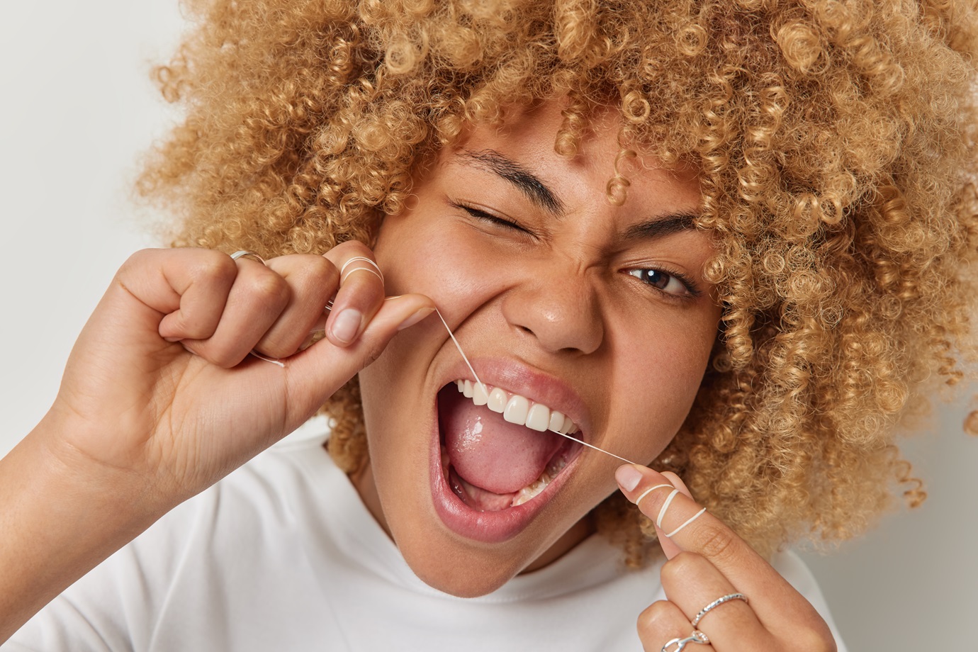 Close up shot of curly haired woman removes food uses thread or dental floss winks eye keeps mouth opened takes care of her mouth cavity poses indoor. Teeth flossing and oral hygiene concept